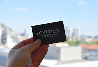 A hand holds a black business card with white text reading 'FORTY TWO Strategy Innovation Design' against a blurred urban skyline backdrop. The sky is clear and blue, suggesting a bright day.