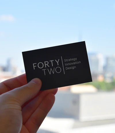 A hand holds a black business card with white text reading 'FORTY TWO Strategy Innovation Design' against a blurred urban skyline backdrop. The sky is clear and blue, suggesting a bright day.