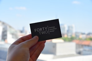 A hand holds a black business card with white text reading 'FORTY TWO Strategy Innovation Design' against a blurred urban skyline backdrop. The sky is clear and blue, suggesting a bright day.