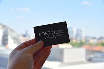A hand holds a black business card with white text reading 'FORTY TWO Strategy Innovation Design' against a blurred urban skyline backdrop. The sky is clear and blue, suggesting a bright day.