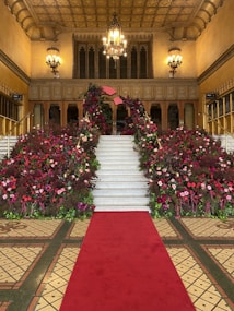 The image displays a grand staircase adorned with lush and vibrant floral arrangements on both sides. The staircase is covered by a red carpet which extends to a mosaic-tiled floor. The background features an ornate interior with decorative chandeliers and large, multi-pane windows. The ceiling has intricate patterns with a golden hue, adding to the opulence of the setting.