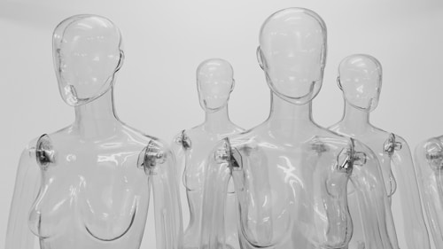Transparent plastic mannequins with smooth, featureless faces stand closely together in an array. The figures have visible joints at the shoulders and elbows, with a glossy surface reflecting light. The background is a plain white setting.