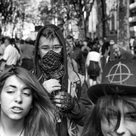 Three young individuals are captured in a crowded outdoor setting. The central figure wears a hood and covers their face with a patterned scarf, exuding a mysterious and rebellious vibe. The person on the left has long hair and a confident expression, while the one on the right wears a hat adorned with an anarchy symbol. The background consists of a bustling crowd and trees lining a street.