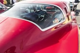 A close-up view of a vintage red car, focusing on its smooth, glossy body and rear window. The interior is visible through the window, showcasing a classic steering wheel and dashboard with various controls. The car is parked outdoors, with blurred figures and other vehicles visible in the background.