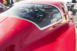 A close-up view of a vintage red car, focusing on its smooth, glossy body and rear window. The interior is visible through the window, showcasing a classic steering wheel and dashboard with various controls. The car is parked outdoors, with blurred figures and other vehicles visible in the background.
