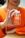 A person is holding a bottle of turmeric and ginger gummies labeled 'Jellybee' with an orange flavor. The person is wearing an orange sweater, and their hands are prominently visible, gently clutching the bottle. The background is slightly blurred.