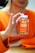 A person is holding a bottle of turmeric and ginger gummies labeled 'Jellybee' with an orange flavor. The person is wearing an orange sweater, and their hands are prominently visible, gently clutching the bottle. The background is slightly blurred.