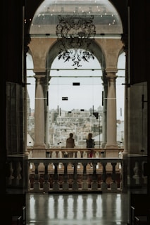 A grand stone archway overlooks a cityscape, with two people standing on a balcony. The elegant arch is adorned with intricate designs. In the foreground, reflective stone flooring leads to the open space, while soft, muted lighting creates a serene atmosphere.