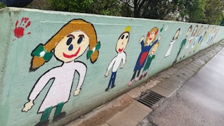 A colorful mural on a concrete wall featuring child-like drawings of smiling people with simple shapes and bright colors. The figures are depicted with exaggerated facial features and colorful clothing, creating a playful and cheerful atmosphere. There are also various handprints in vibrant colors along the wall.