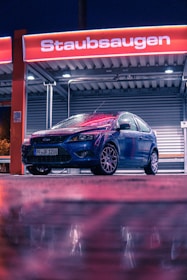 A blue car is parked under a red sign at a car wash station at night. The ground and the car are wet, likely due to rain. The environment is illuminated by artificial lights creating reflections on the wet surface.