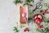A marble countertop is adorned with a parchment-lined container of freshly made pink ice cream next to a scoop. There are scattered strawberries, a bowl filled with scoops of the ice cream topped with strawberries, an open bottle of syrup, and large vintage scissors. Delicate white flowers and sprigs of greenery are arranged around the setup.