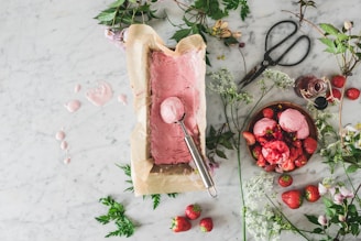 A marble countertop is adorned with a parchment-lined container of freshly made pink ice cream next to a scoop. There are scattered strawberries, a bowl filled with scoops of the ice cream topped with strawberries, an open bottle of syrup, and large vintage scissors. Delicate white flowers and sprigs of greenery are arranged around the setup.