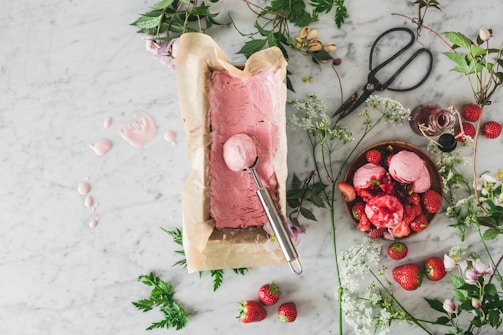 A marble countertop is adorned with a parchment-lined container of freshly made pink ice cream next to a scoop. There are scattered strawberries, a bowl filled with scoops of the ice cream topped with strawberries, an open bottle of syrup, and large vintage scissors. Delicate white flowers and sprigs of greenery are arranged around the setup.