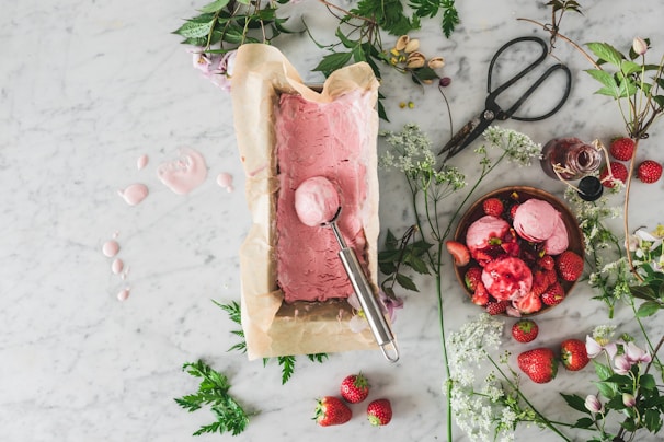 A marble countertop is adorned with a parchment-lined container of freshly made pink ice cream next to a scoop. There are scattered strawberries, a bowl filled with scoops of the ice cream topped with strawberries, an open bottle of syrup, and large vintage scissors. Delicate white flowers and sprigs of greenery are arranged around the setup.