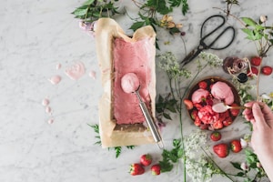 A rustic and artistic food scene featuring freshly made pink ice cream being scooped from a parchment-lined container on a marble surface. The setup is surrounded by fresh strawberries, green leaves, flowering branches, and a pair of vintage scissors. A wooden bowl holds scoops of strawberry ice cream garnished with strawberries, with a hand serving the dessert with a spoon.