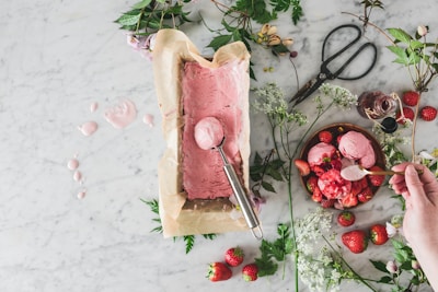A rustic and artistic food scene featuring freshly made pink ice cream being scooped from a parchment-lined container on a marble surface. The setup is surrounded by fresh strawberries, green leaves, flowering branches, and a pair of vintage scissors. A wooden bowl holds scoops of strawberry ice cream garnished with strawberries, with a hand serving the dessert with a spoon.