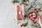 A rustic and artistic food scene featuring freshly made pink ice cream being scooped from a parchment-lined container on a marble surface. The setup is surrounded by fresh strawberries, green leaves, flowering branches, and a pair of vintage scissors. A wooden bowl holds scoops of strawberry ice cream garnished with strawberries, with a hand serving the dessert with a spoon.