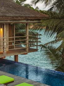A tranquil scene of a wooden cabin with a thatched roof overlooking an infinity pool. The setting is surrounded by lush palm trees with a view out to a calm, blue sea. The warm glow from a lamp inside the cabin adds a cozy ambiance.