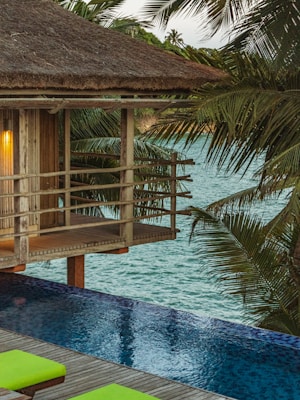A tranquil scene of a wooden cabin with a thatched roof overlooking an infinity pool. The setting is surrounded by lush palm trees with a view out to a calm, blue sea. The warm glow from a lamp inside the cabin adds a cozy ambiance.
