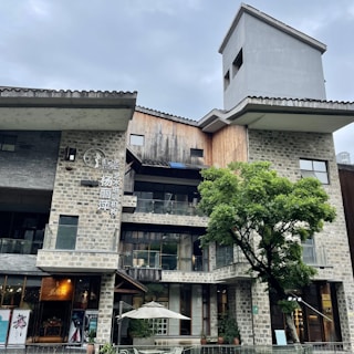 An architectural structure with a modern design, featuring a mix of stone and wooden materials. The building has multiple levels with large glass windows. A prominent tree is in front of the building, and there's an outdoor area with a white umbrella over a seating area. The sky is overcast.