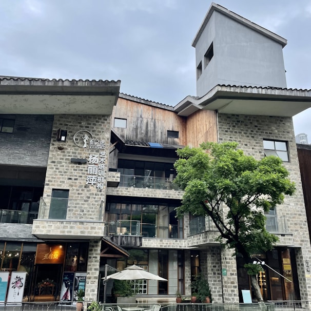 An architectural structure with a modern design, featuring a mix of stone and wooden materials. The building has multiple levels with large glass windows. A prominent tree is in front of the building, and there's an outdoor area with a white umbrella over a seating area. The sky is overcast.