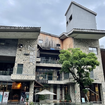 An architectural structure with a modern design, featuring a mix of stone and wooden materials. The building has multiple levels with large glass windows. A prominent tree is in front of the building, and there's an outdoor area with a white umbrella over a seating area. The sky is overcast.