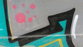 Vibrant graffiti art featuring bold geometric shapes in black with colorful accents. A background of grey is adorned with red circular patterns. Vivid teal and yellow elements add contrast and playfulness to the composition, which is painted on a textured wall surface.
