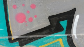 Vibrant graffiti art featuring bold geometric shapes in black with colorful accents. A background of grey is adorned with red circular patterns. Vivid teal and yellow elements add contrast and playfulness to the composition, which is painted on a textured wall surface.