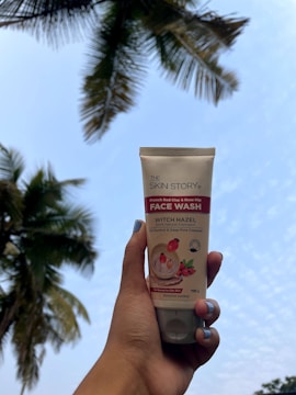 A hand with painted nails holds a tube of face wash with a background of a partly cloudy sky and palm tree leaves.
