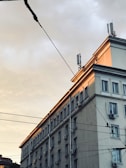 A multi-story urban building with numerous windows and air conditioning units on the exterior walls. The upper part of the building is illuminated by the soft, warm light of a setting or rising sun. Several utility wires are visible in the foreground, stretching across the scene.