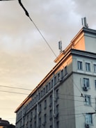 A multi-story urban building with numerous windows and air conditioning units on the exterior walls. The upper part of the building is illuminated by the soft, warm light of a setting or rising sun. Several utility wires are visible in the foreground, stretching across the scene.