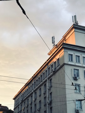 A multi-story urban building with numerous windows and air conditioning units on the exterior walls. The upper part of the building is illuminated by the soft, warm light of a setting or rising sun. Several utility wires are visible in the foreground, stretching across the scene.