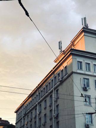 A multi-story urban building with numerous windows and air conditioning units on the exterior walls. The upper part of the building is illuminated by the soft, warm light of a setting or rising sun. Several utility wires are visible in the foreground, stretching across the scene.