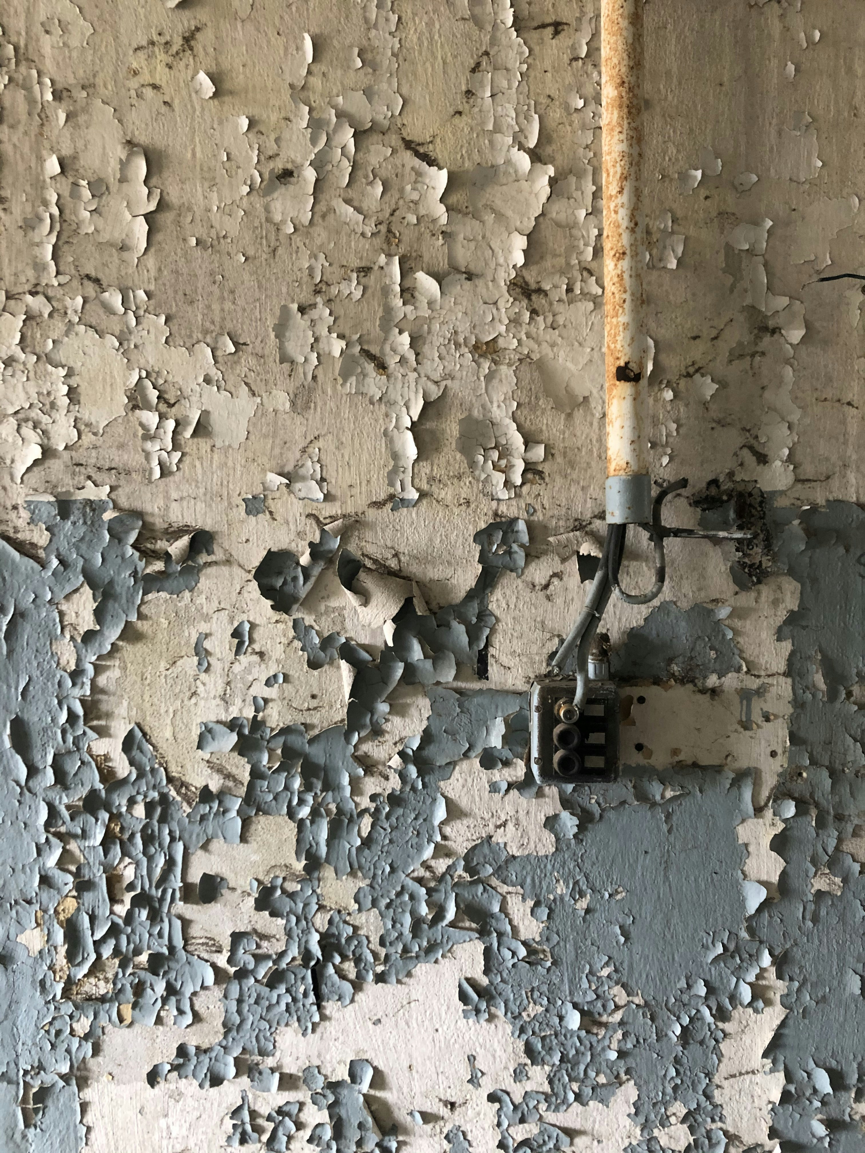 Peeling paint covers the surface of an old wall, revealing multiple layers of faded and chipped coatings. A rusted metal pipe runs vertically along the wall, connected to a vintage electrical box.