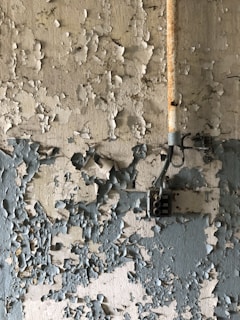 Peeling paint covers the surface of an old wall, revealing multiple layers of faded and chipped coatings. A rusted metal pipe runs vertically along the wall, connected to a vintage electrical box.