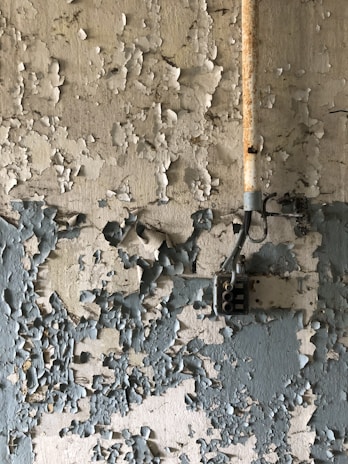 Peeling paint covers the surface of an old wall, revealing multiple layers of faded and chipped coatings. A rusted metal pipe runs vertically along the wall, connected to a vintage electrical box.