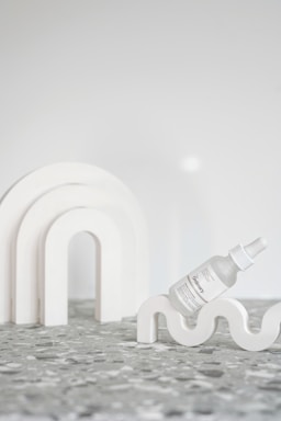 A minimalist arrangement featuring a bottle of skincare serum with a dropper, labeled 'the ordinary', resting on a wavy white decorative object. Behind it, an arch-shaped sculpture complements the aesthetic. The surface is a terrazzo-style texture in gray tones, and the background is a plain, soft white.