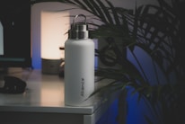 A white insulated water bottle with a metallic lid sits on a desk, next to a lamp that emits a soft glow. The word 'Alliance' is printed vertically on the bottle. There are large leafy plants present, casting shadows on the wall, creating a calm and minimalistic atmosphere.