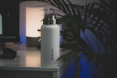 A white insulated water bottle with a metallic lid sits on a desk, next to a lamp that emits a soft glow. The word 'Alliance' is printed vertically on the bottle. There are large leafy plants present, casting shadows on the wall, creating a calm and minimalistic atmosphere.