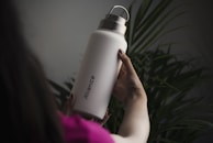 A person is holding a large, white water bottle with a metal cap. The word 'Alliance' is printed vertically on the bottle. The background is filled with green plant leaves, creating a natural setting. The person appears to be wearing a pink garment, and the lighting is dim, giving a soft and calm atmosphere.