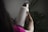 A person is holding a large, white water bottle with a metal cap. The word 'Alliance' is printed vertically on the bottle. The background is filled with green plant leaves, creating a natural setting. The person appears to be wearing a pink garment, and the lighting is dim, giving a soft and calm atmosphere.