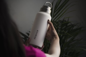 A person is holding a large, white water bottle with a metal cap. The word 'Alliance' is printed vertically on the bottle. The background is filled with green plant leaves, creating a natural setting. The person appears to be wearing a pink garment, and the lighting is dim, giving a soft and calm atmosphere.