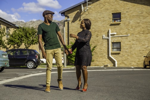A man and a woman are walking together in an outdoor residential area. The man is wearing a green shirt, beige pants, and a cap, while the woman is dressed in a black dress and red high heels. They are holding hands and appear to be engaged in conversation. The background features a brick building, parked cars, and greenery under a clear sky with some clouds.