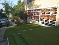 A well-maintained lawn area with green artificial grass is shown. There are steps covered with the same grass leading up to a building with a brick facade, which has several rectangular flower pots arranged on vertical racks attached to the wall. These pots contain red and pink flowers. To the left, there is a small tree and to the far left, parked cars are visible next to a concrete path.