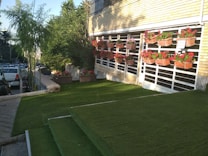 A well-maintained lawn area with green artificial grass is shown. There are steps covered with the same grass leading up to a building with a brick facade, which has several rectangular flower pots arranged on vertical racks attached to the wall. These pots contain red and pink flowers. To the left, there is a small tree and to the far left, parked cars are visible next to a concrete path.