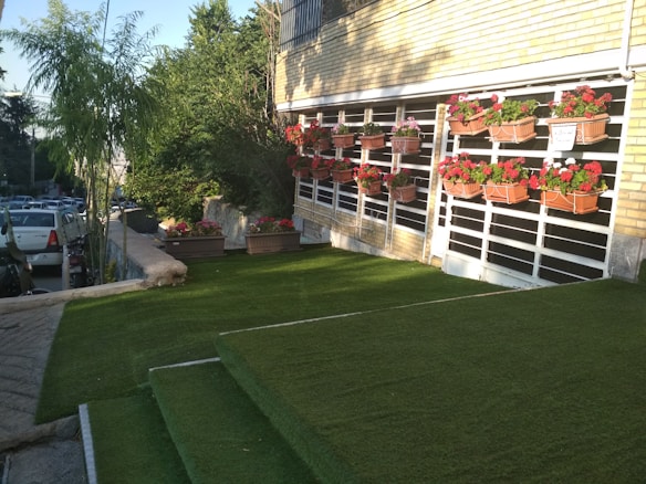 A well-maintained lawn area with green artificial grass is shown. There are steps covered with the same grass leading up to a building with a brick facade, which has several rectangular flower pots arranged on vertical racks attached to the wall. These pots contain red and pink flowers. To the left, there is a small tree and to the far left, parked cars are visible next to a concrete path.