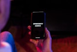 A hand is holding a black smartphone with the screen displaying the text 'OPPORTUNITY KNOCKS' in white font. The phone is surrounded by a dimly lit environment with hints of red and blue lighting.