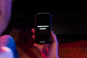 A hand is holding a black smartphone with the screen displaying the text 'OPPORTUNITY KNOCKS' in white font. The phone is surrounded by a dimly lit environment with hints of red and blue lighting.