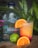A bottle of silver tequila labeled 'Ghost Pepper' is placed next to a tropical cocktail garnished with a slice of orange. A whole lime and a sectioned orange are on the wooden surface in front. The background includes green foliage and small pink flowers, creating a fresh, natural setting.