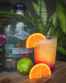 A bottle of silver tequila labeled 'Ghost Pepper' is placed next to a tropical cocktail garnished with a slice of orange. A whole lime and a sectioned orange are on the wooden surface in front. The background includes green foliage and small pink flowers, creating a fresh, natural setting.